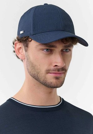 Mann mit kurzem Bart, der eine strukturierte marineblaue Baseballkappe und einen marineblauen Pullover mit weiß abgesetztem Kragen trägt, schaut nach rechts vor weißem Hintergrund.