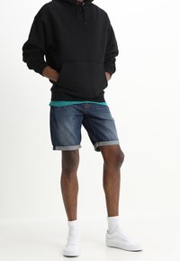 Persona con sudadera negra, camiseta verde azulado, pantalones cortos de denim arremangados y zapatillas blancas de pie con las manos en los bolsillos.