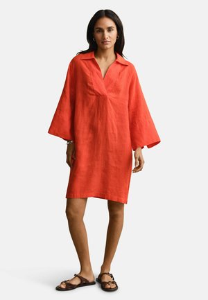 Femme portant une robe rouge ample à manches longues, longueur au genou, avec un col et un décolleté en V, associée à des sandales plates marron foncé.