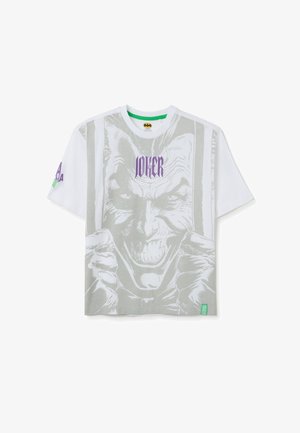 T-shirt bianco a maniche corte con una grande grafica grigia del Joker sulla parte anteriore. Testo "JOKER" in viola sopra l'immagine. Etichetta di branding verde.