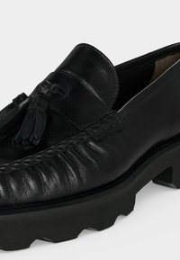 Mocassin noir en cuir avec glands, détails cousus et semelle épaisse à rainures, montré en gros plan sur fond blanc.
