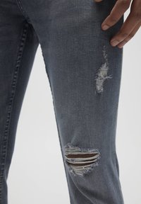 Jeans de mezclilla azul oscuro con desgaste en la rodilla, bordes deshilachados y un desgarro horizontal que revela una tela marrón debajo.