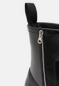 Botas de cuero negras con cremallera lateral y un tirador plateado, además de un bucle de tela en la parte superior para facilitar su uso. Textura suave.