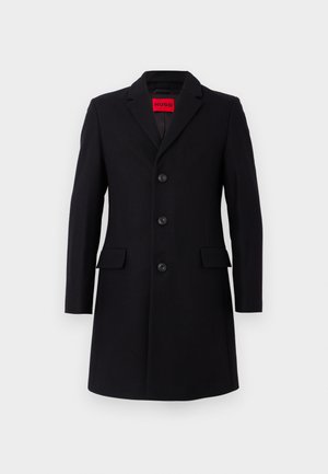 Cappotto in lana nera con vestibilità sartoriale. Presenta un colletto a punta, cinque bottoni e due tasche frontali. Interno foderato. Logo rosso visibile.