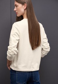 Sudadera de color beige claro con puños y dobladillo acanalados, que presenta un corte relajado. La parte posterior es lisa con una sutil textura, combinada con unos jeans de color azul oscuro.