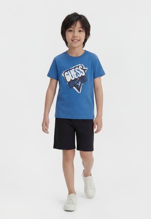 Dreng går fremad iført en blå t-shirt med grafik, sorte shorts og hvide sneakers mod en ensfarvet hvid baggrund.