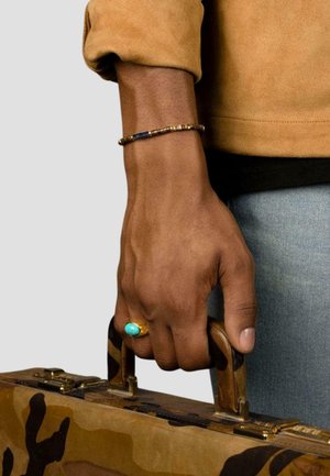 Bracelet with blue and gold beads on wrist; hand gripping a camo-patterned briefcase; gold ring with a turquoise stone.