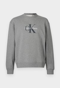 Šedá mikina s okrúhlym výstrihom a rebrovanými manžetami, so sieťotlačovým logom „Calvin Klein“ v kontrastných farbách na prednej strane.