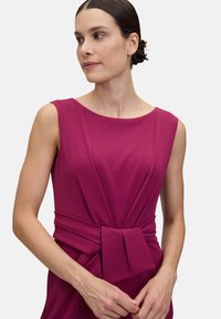 Fuchsiaklänning med båtformad halsringning och ärmar med kap. Har veckad detalj och en knuten rosett i midjan. Slät tygtextur.