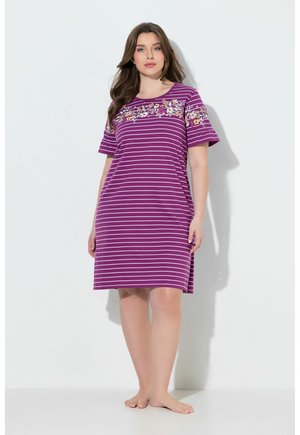 Femme portant une robe rayée violette et blanche mi-longue avec une broderie florale sur la poitrine et les manches, debout pieds nus contre un mur blanc.