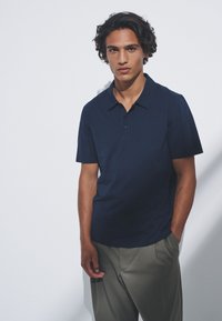 Mornarsko modra polo majica s klasičnim ovratnikom, kratkimi rokavi in dvojnim gumbom na sprednji strani, kombinirana s svetlo sivimi hlačami. Gladek tekstura.