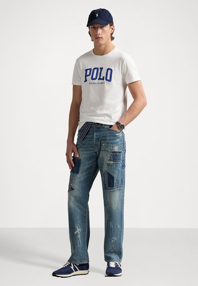 Tricou alb din bumbac cu logo-ul albastru "POLO", blugi deteriorați cu aplicații, șapcă bleumarin și adidași blue. Modelul stă în picioare cu mâinile relaxate.