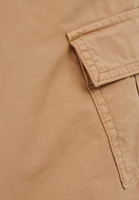 Khaki stof met een gladde textuur. Beschikt over een zak met een flap en dubbel gestikte randen. Subtiele glans.