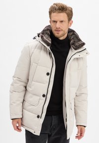 Man in een lichtbeige gewatteerde winterjas met een bontgevoerde kraag over een zwarte coltrui, staande voor een effen achtergrond.