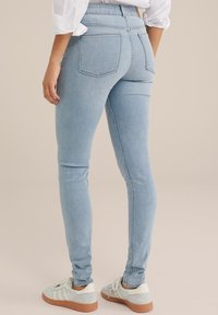 Hellblaue Skinny-Jeans mit einer glatten Denim-Oberfläche, hoher Taille und zwei Gesäßtaschen, kombiniert mit grauen Sneakers mit weißen Streifen.