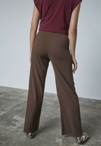 Bruna, vida byxor i mjukt tyg, parat med svarta och vita mönstrade ballerina-skor. Modellens topp är vinröd med korta ärmar.