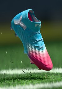 Crampons de football bleu et rose avec des lacets et des crampons, soulevant de l'herbe et positionnés au-dessus d'une ligne blanche sur un terrain vert.