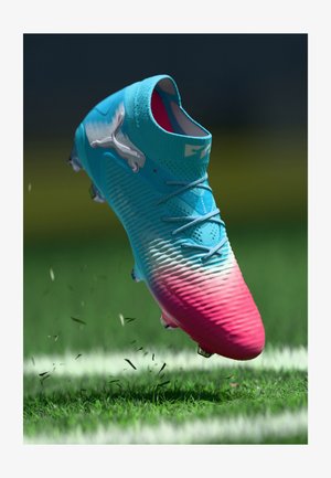 Chuteira de futebol azul e rosa com atacadores e cravos, levantando relva e posicionada acima de uma linha branca num campo verde.