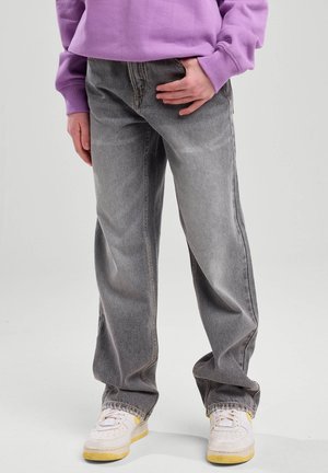 Persoon draagt losse grijze jeans, witte sneakers met gele zolen en een paarse trui, staande tegen een effen lichte achtergrond.