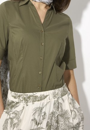 Frau trägt ein olivgrünes, geknöpftes Hemd mit hochgekrempelten Ärmeln und einen weißen Rock mit detailliertem tropischem Vogel- und Pflanzenmuster.