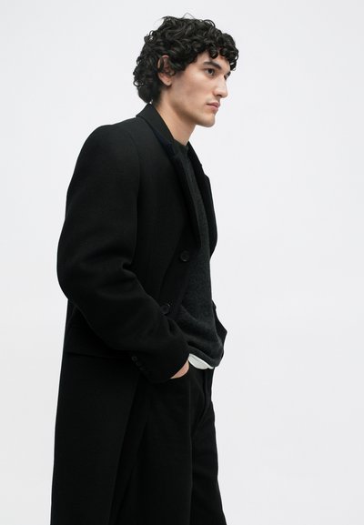 Άνδρας με σγουρά μαλλιά που φοράει ένα μαύρο παλτό και σκοτεινό πουλόβερ, στέκεται προφίλ με τα χέρια στις τσέπες, σε ένα απλό ανοιχτό φόντο.