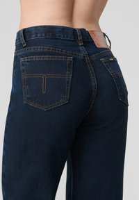 Sötétkék farmernadrág sima textúrával, látható varrással és öt zsebbel. Bőr címke található a hátsó derékrészen.