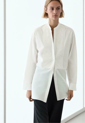 Ung person med kort blondt hår iført en hvid skjorte med knapper og broderede frontpaneler samt sorte bukser, stående indendørs.