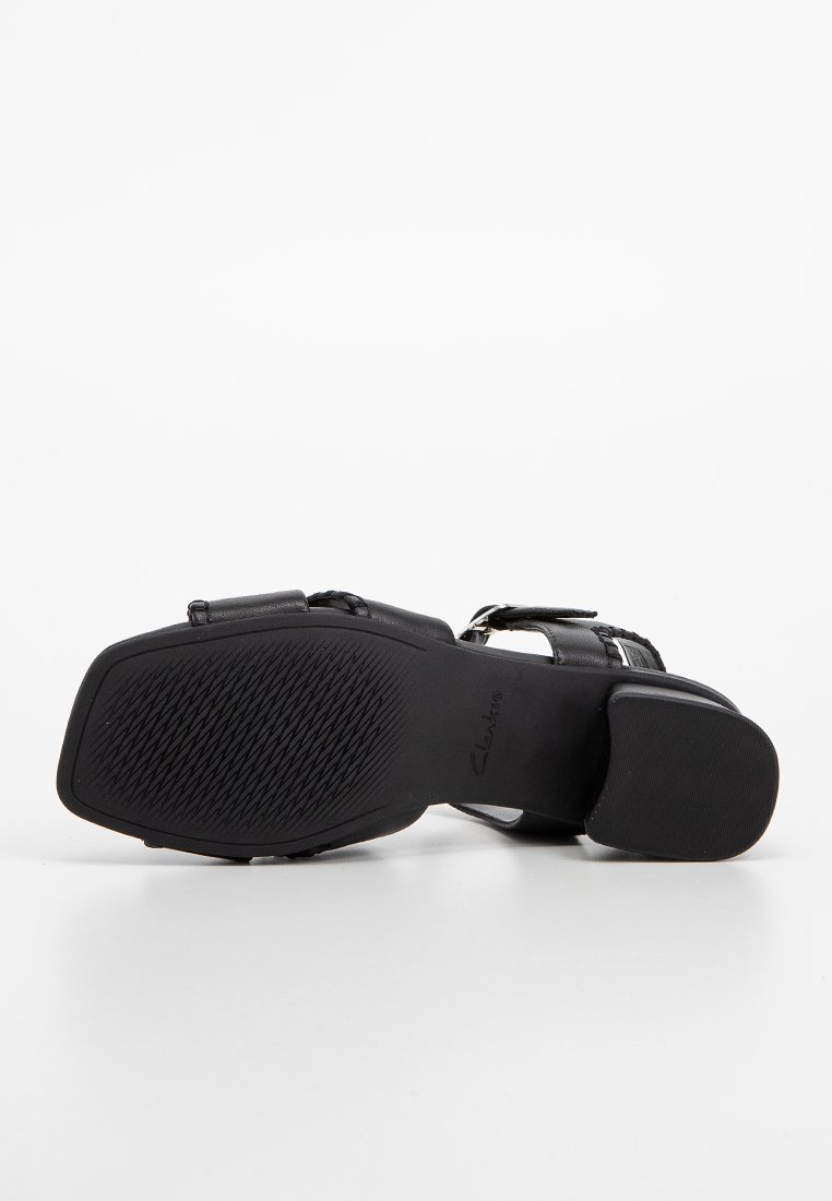 Sandalia negra con tacón cuadrado y suela texturizada, con correa de hebilla, vista desde abajo sobre un fondo blanco.