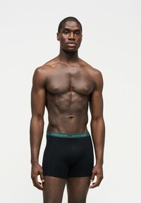 Μυώδης άνδρας που στέκεται με χαλαρά χέρια, φορώντας μαύρα ανδρικά εσώρουχα Björn Borg με πράσινη ζώνη μέσης σε απλό λευκό φόντο.