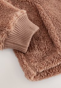 Morbido tessuto in pile marrone soffice con polsino a costine, che mostra il contrasto di texture tra materiali morbidi e lisci.