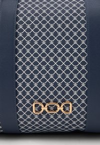 Kék táska, geometrikus fehér mintával díszített anyaggal, bőr kiegészítőkkel és arany lánc logóval az alján.