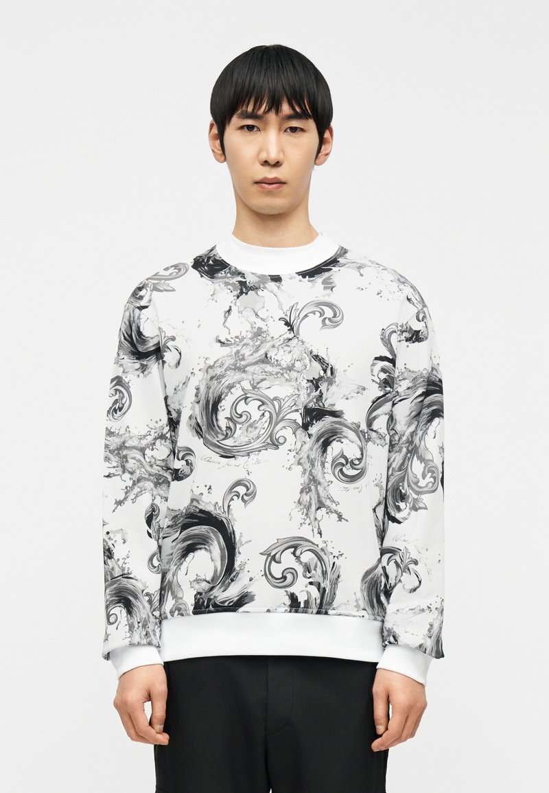 Άνδρας με κοντά μαύρα μαλλιά που φορεί λευκό φούτερ με μαύρα και γκρι αφηρημένα στροβιλώδη σχέδια, συνδυασμένο με μαύρο παντελόνι, στέκεται μπροστά σε λευκό φόντο.