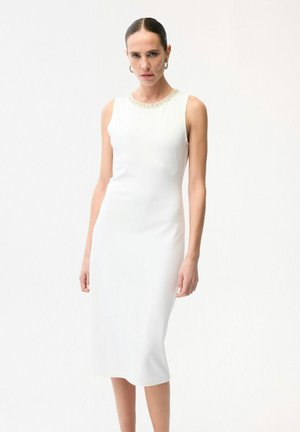 Vrouw draagt een mouwloze witte midijurk met een met kralen versierde halslijn, staand tegen een effen witte achtergrond.