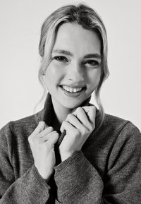 Maglione grigio a maglia con colletto a risvolto, caratterizzato da una texture morbida. Il modello tiene il colletto con entrambe le mani, sorridendo, su uno sfondo semplice.