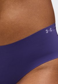 Κοντινό πλάνο στο ισχίο ενός ατόμου που φοράει μοβ αθλητικά σορτς Under Armour χωρίς ραφές, με λογότυπο στη ζώνη.