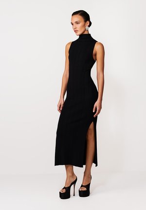 Czarna, prążkowana sukienka maxi z wysokim kołnierzem, bocznym rozcięciem i dopasowaną sylwetką. Noszona z czarnymi platformowymi szpilkami, prezentująca elegancki, nowoczesny styl.