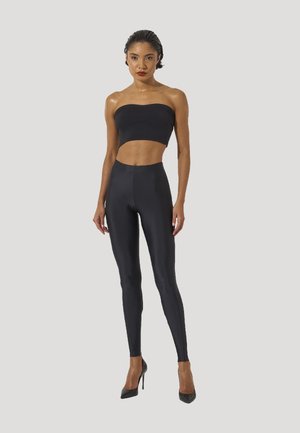 Kvinde står iført en sort stropløs crop top, sorte, højtaljede glansfulde leggings og sorte højhælede sko mod en ensfarvet baggrund.