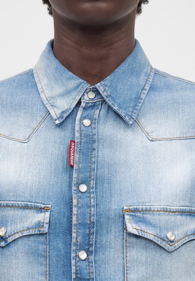 Κοντινό πλάνο ενός ατόμου που φοράει ανοιχτό μπλε τζιν πουκάμισο με κουμπιά τύπου κλικ και δύο τσέπες στο στήθος, με κόκκινη ετικέτα DSQUARED2.