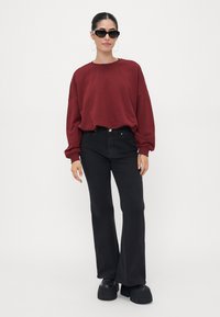 Sudadera oversized burdeos, jeans negros acampanados, zapatos negros de plataforma y gafas de sol oscuras, contra un fondo blanco liso.