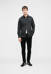 Fekete hétköznapi ing két mellzsebbel, gombos elülső résszel, fekete slim-fit nadrággal és fekete, fehér részletekkel díszített tornacipővel párosítva.