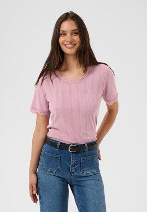 Smilende kvinde med langt brunt hår iført en pink top med korte ærmer og blonderkant samt blå jeans med lomme foran og sort bælte.