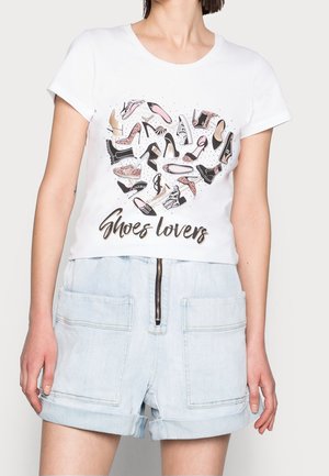Valkoinen T-paita, jossa erilaisia naisten kenkäkuvituksia sydämen muotoon järjestettynä tekstin "Shoes lovers" yläpuolella, yhdistetty vaaleisiin farkkushortseihin.