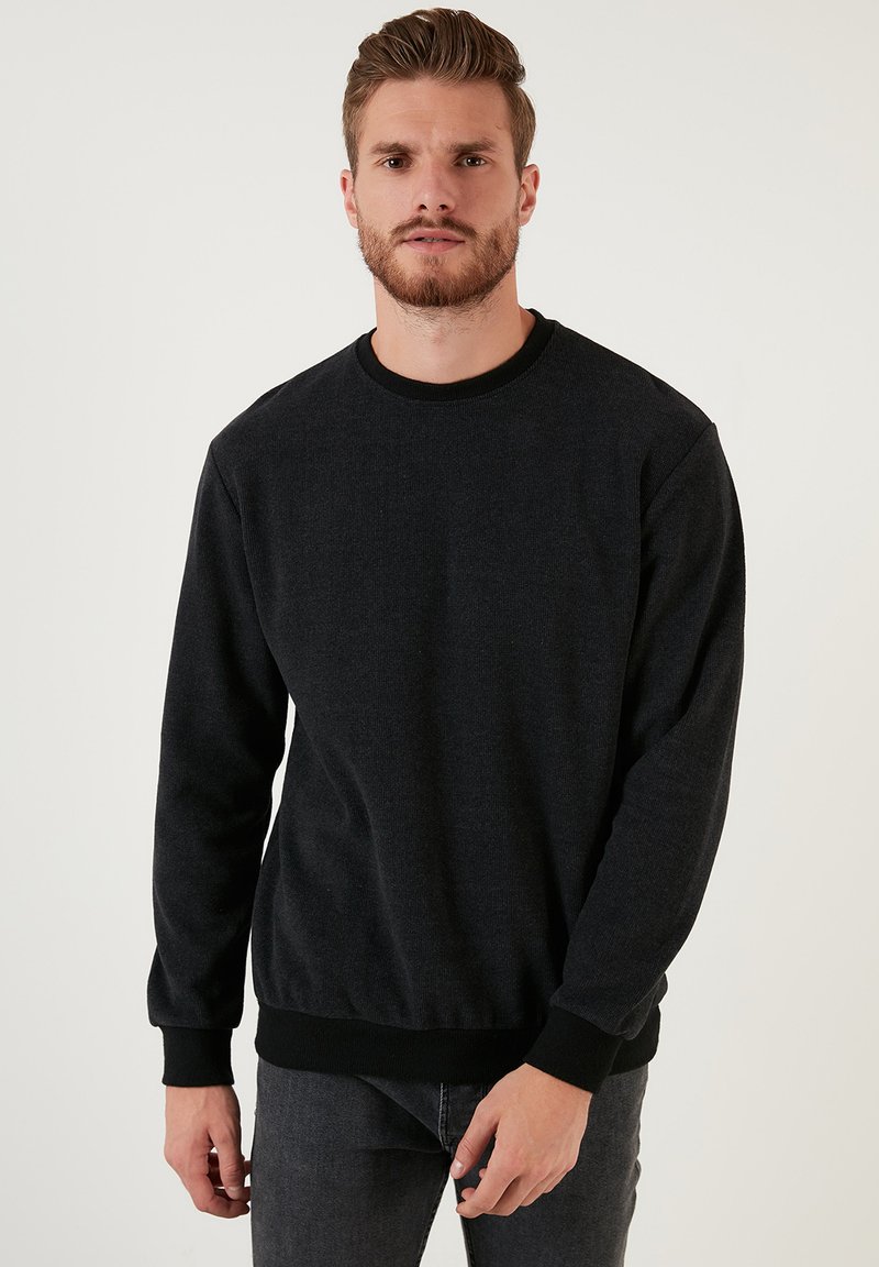 Schwarzer Sweatshirt aus strukturiertem Stoff mit Rundhalsausschnitt und gerippten Bündchen. Das Model trägt es mit einer schmal geschnittenen dunkelblauen Jeans.