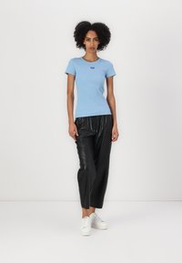Camiseta de manga corta azul claro con cuello redondo y un pequeño logo, combinada con pantalones de cuero negros y zapatillas blancas.