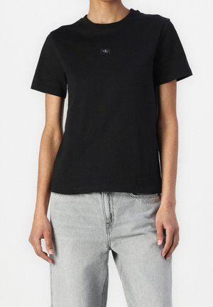 Black cotton t-shirt with short sleeves and a crew neckline, featuring a small logo patch on the chest and a straight hem. Worn with gray jeans.