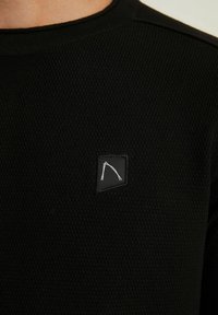 Černý svetr s texturou, na levé straně malá čtvercová nášivka s bílým motivem. Zesílené švy na ramenou.