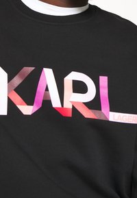 Camiseta negra con un gran logo "KARL" en tonos degradados de rosa a púrpura. Cuello clásico de crew con ribete blanco en el interior.