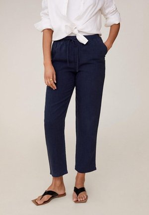 Woman wearing navy blue drawstring pants, white knotted long-sleeve shirt, and black thong sandals standing with one hand in pocket.