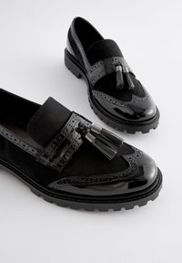Schwarze Loafers aus einer Kombination von Wildleder und Lackleder. Mit Quasten, Brogue-Details und einer dicken Profilsohle für besseren Halt.