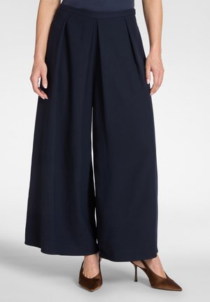 Frau trägt marineblaue, weit geschnittene Hose und braune, spitz zulaufende High Heels, steht vor einem schlichten Hintergrund.